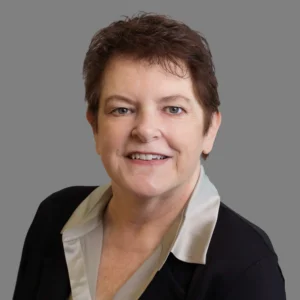 A middle-aged woman with short brown hair, wearing a black blazer and a light-colored blouse, smiling in front of a plain gray background.