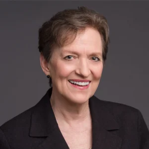 A smiling older woman with short brown hair, wearing a dark blazer, posed against a plain gray background.