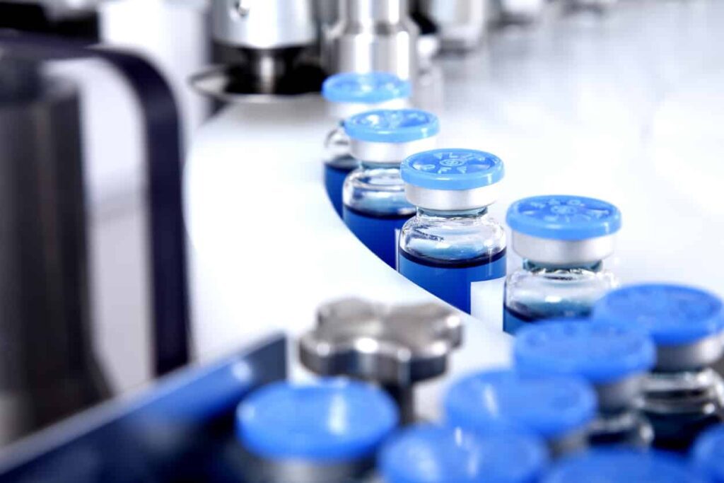 Close-up of several glass vials with blue caps filled with liquid, moving along a conveyor belt in a pharmaceutical manufacturing facility.