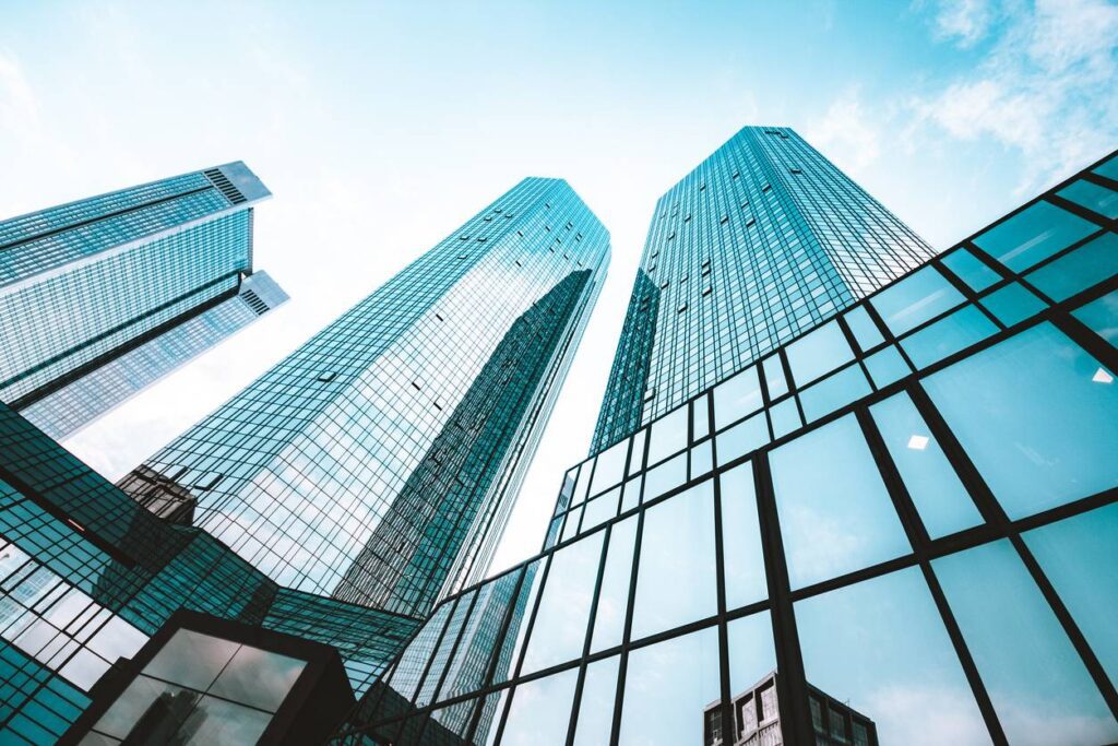 Tall glass skyscrapers with reflective windows rise into a blue sky, viewed from a low angle.