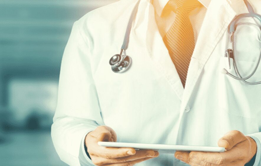 A doctor wearing a white coat and stethoscope holds a tablet device, with a blurred background suggesting a medical setting.