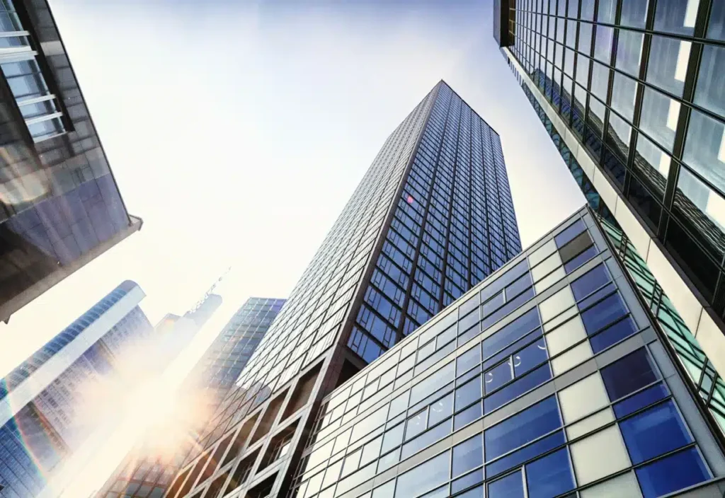 Tall glass office buildings viewed from below with sunlight reflecting off the windows and a clear sky in the background.