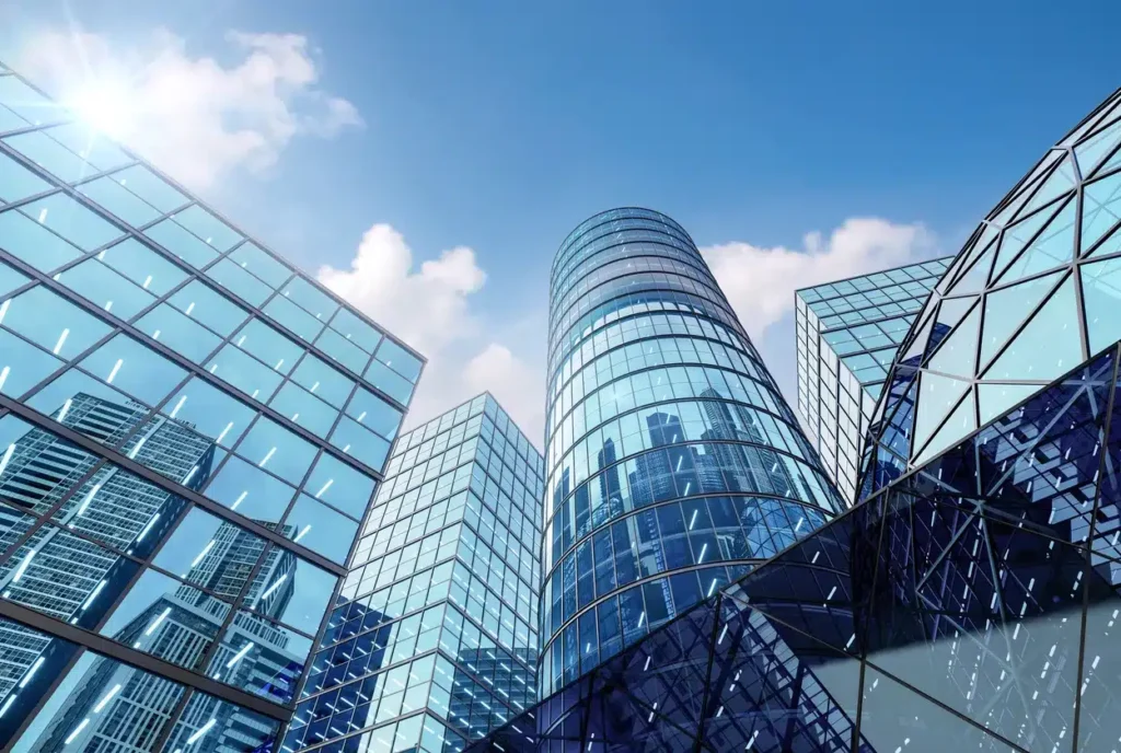 Modern glass skyscrapers against a blue sky with scattered clouds, reflecting sunlight and surrounding buildings.