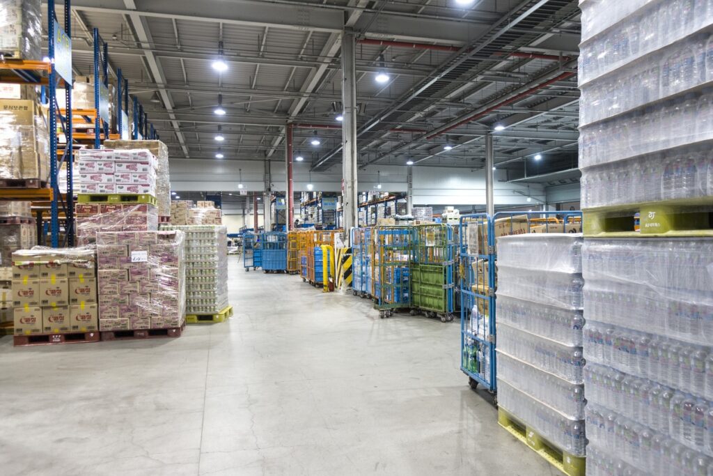Interior of a warehouse with shelves and pallets stacked with goods, including bottled water and boxes, under bright overhead lighting.