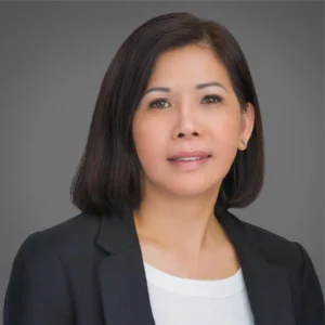 A woman with straight dark hair, wearing a black blazer and white top, poses against a plain gray background.