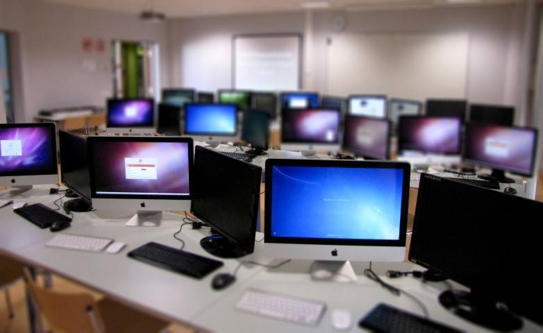 A computer lab with multiple desktop monitors and computers arranged on tables, some screens are on and displaying login or desktop pages.