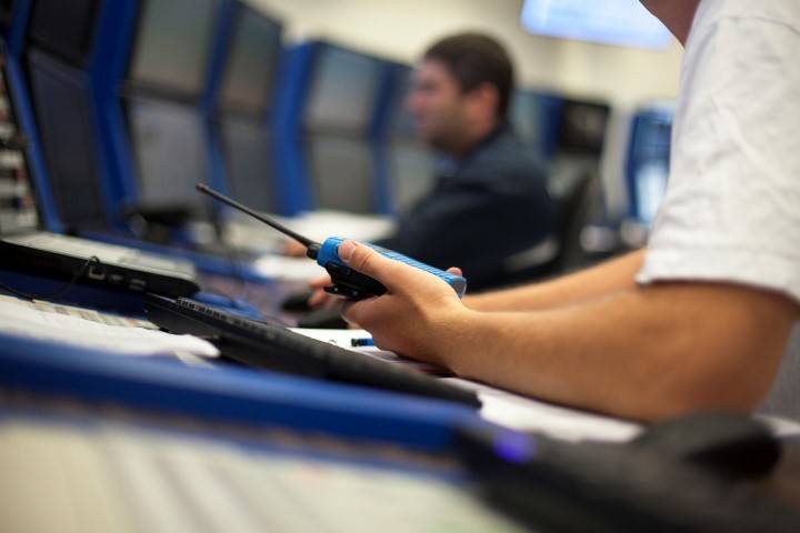 A person holds a walkie-talkie at a desk with computer monitors, while another person works in the background.