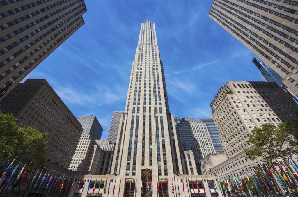 Tall art deco skyscraper at Rockefeller Center in New York City, surrounded by other buildings and flags from various countries, under a blue sky.