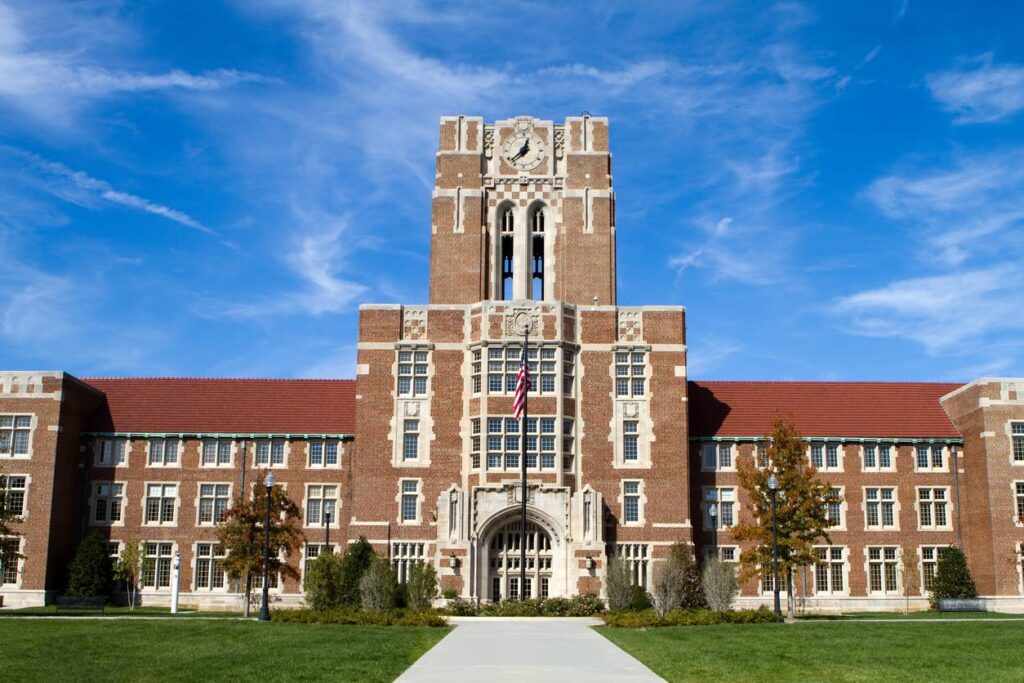 A large brick academic building with a clock tower, arched entrance, and an American flag, set against a blue sky with scattered clouds.