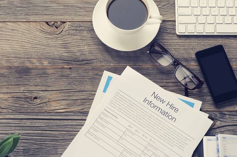New hire information forms, eyeglasses, a smartphone, a keyboard, and a cup of coffee on a wooden desk.