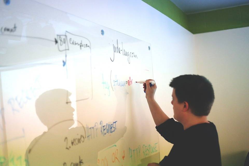 A person writes on a whiteboard with a red marker in an office setting, with various diagrams and text already on the board.