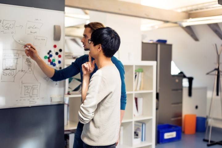 Two people stand at a whiteboard discussing diagrams and notes in a modern office setting, with shelves and storage in the background.