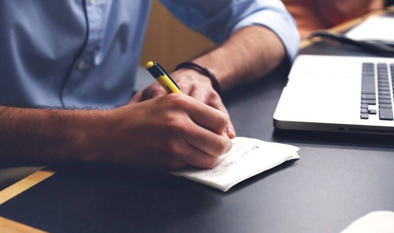 A person writes notes with a pen on a piece of paper next to a laptop on a desk.