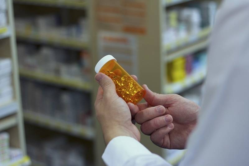 A person in a white coat holds an orange prescription pill bottle in a pharmacy setting, with shelves of medication in the background.