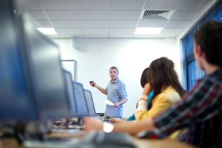 A man is standing and teaching in front of a classroom, gesturing toward a whiteboard, while students sit at computers and listen.