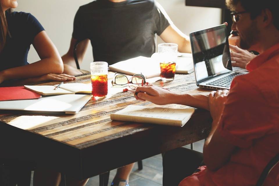 Four people sit around a wooden table with notebooks, a laptop, and drinks, appearing to have a meeting or discussion in a casual setting.