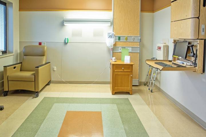A hospital room with a beige reclining chair, wooden cabinet, medical equipment, and a fold-out wall desk, featuring neutral-colored tiled floors and light-colored walls.