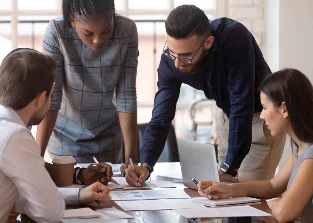 Four people sit and stand around a table, discussing documents and charts, with a laptop and coffee cup visible on the desk.