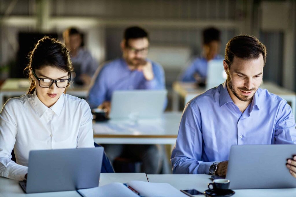 Several people work on laptops in an office setting, with two individuals in the foreground focused on their screens as they engage with innovative training solutions.