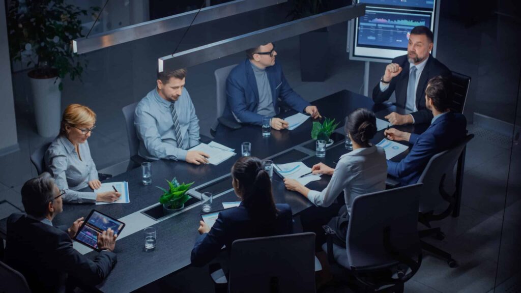 Eight people in business attire sit around a conference table in an office, engaged in a meeting with laptops, tablets, and documents.