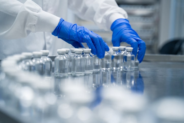 A person wearing blue gloves and a white lab coat arranges rows of clear glass vials with white caps on a stainless steel surface in a laboratory setting.