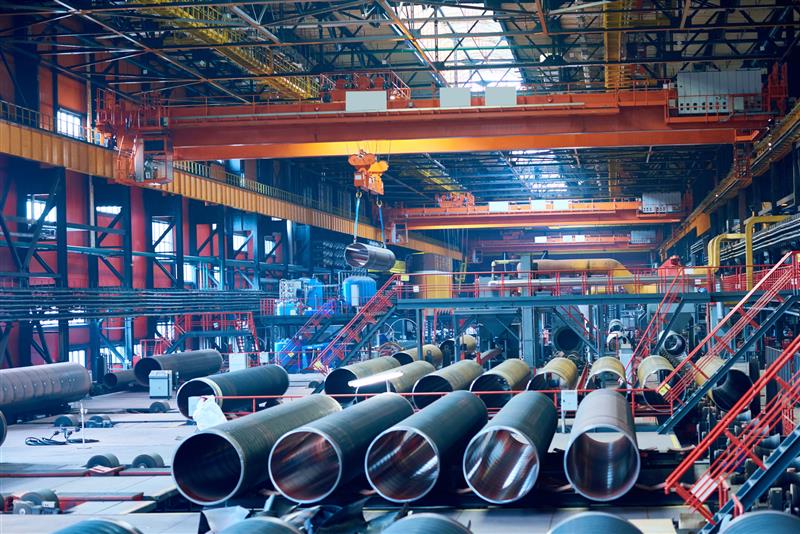 Large industrial factory floor with numerous steel pipes, machinery, cranes, and workers assembling or moving metal components under bright lighting—an ideal environment for implementing advanced Training Solutions.