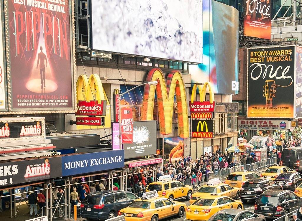 Crowded street scene in Times Square, New York City, with yellow taxis, people in line, McDonald's signs, and large Broadway show billboards.