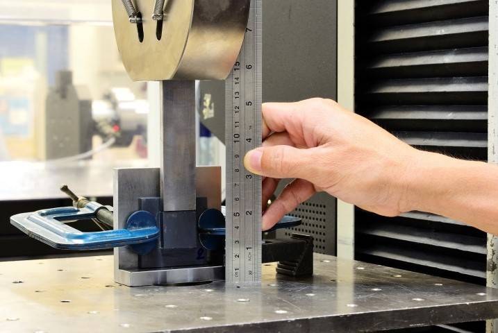 A hand holds a metal ruler to measure the height of a clamped object on a metal workbench, likely in a laboratory or workshop setting.