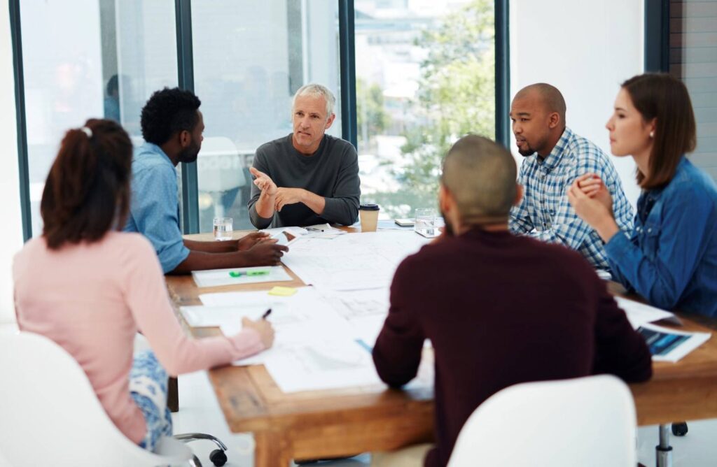 Six people sit around a table in a brightly lit office, engaged in discussion with papers, notebooks, and pens spread out in front of them.