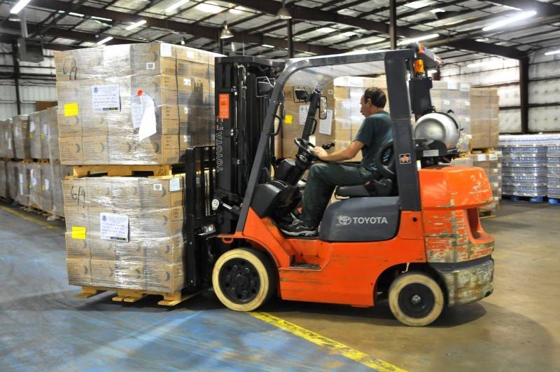 A man operates an orange forklift to move stacked pallets of boxed goods inside a warehouse.