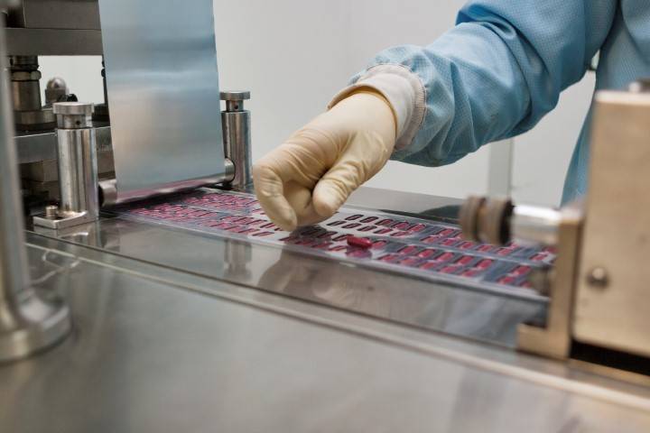 A person wearing gloves and a blue lab coat inspects or processes blister packs of red pills on a pharmaceutical production line.