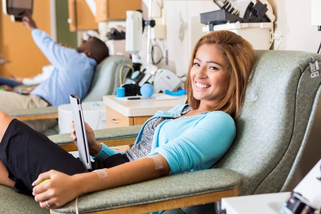 Woman sitting in a medical chair and smiling at the camera after donating blood, holding a clipboard; another person is in the background.