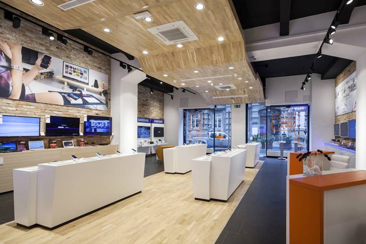 Modern electronics store interior with wooden flooring, white display tables, wall-mounted screens, and large windows facing the street.