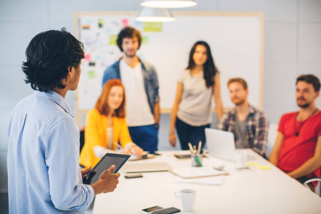A person with a tablet presents to a group of five people sitting and standing around a table in a meeting room with a whiteboard.