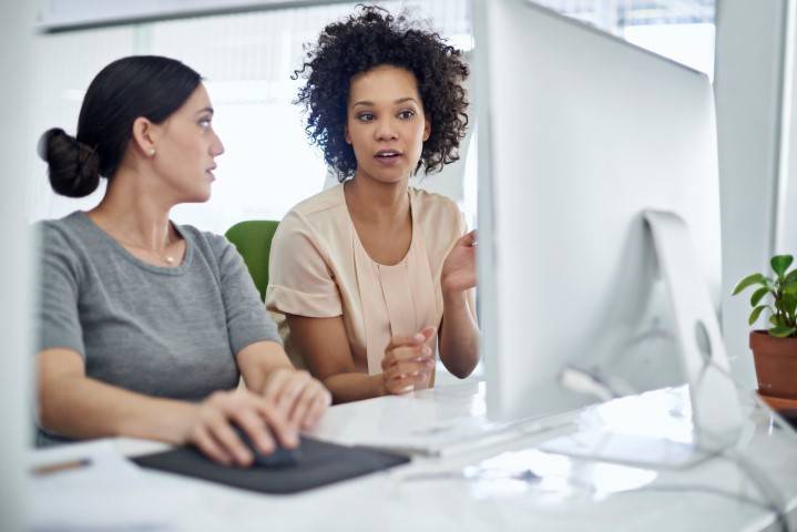 Two women sit at a desk working on a computer, engaged in conversation. One uses the keyboard and mouse while the other gestures as she speaks.