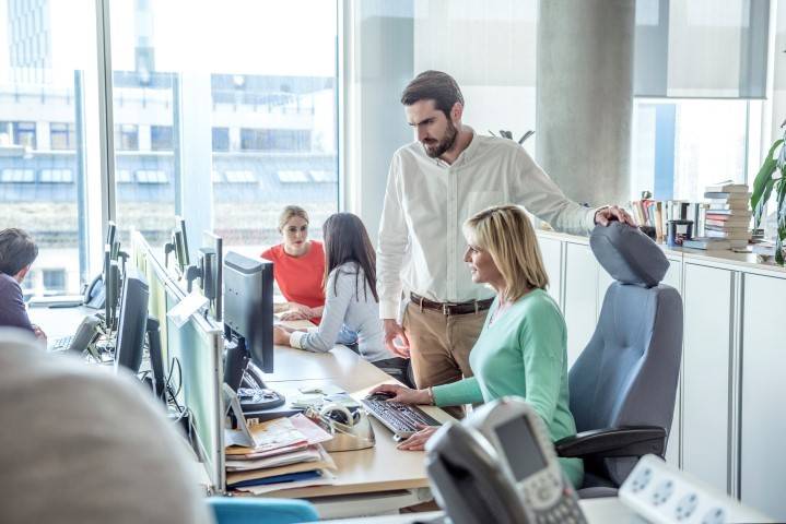 Five people work at computer desks in a modern office with large windows. One man stands beside a seated woman, both looking at a computer monitor.