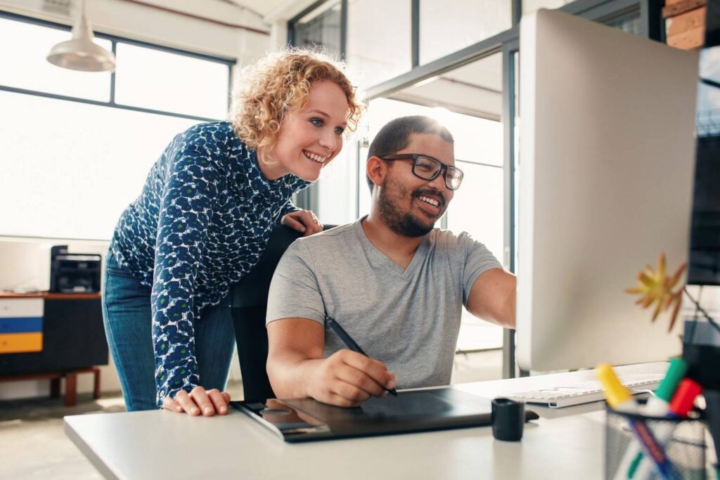 Two people in an office, one sitting at a desk using a computer and graphics tablet, the other standing beside and looking at the screen, both smiling.