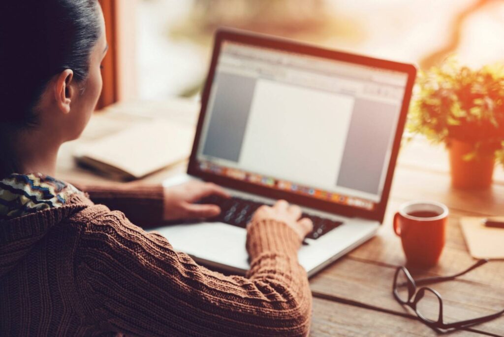 Person wearing a brown sweater typing on a laptop at a wooden desk with a cup of coffee, eyeglasses, a notebook, and a small potted plant nearby.
