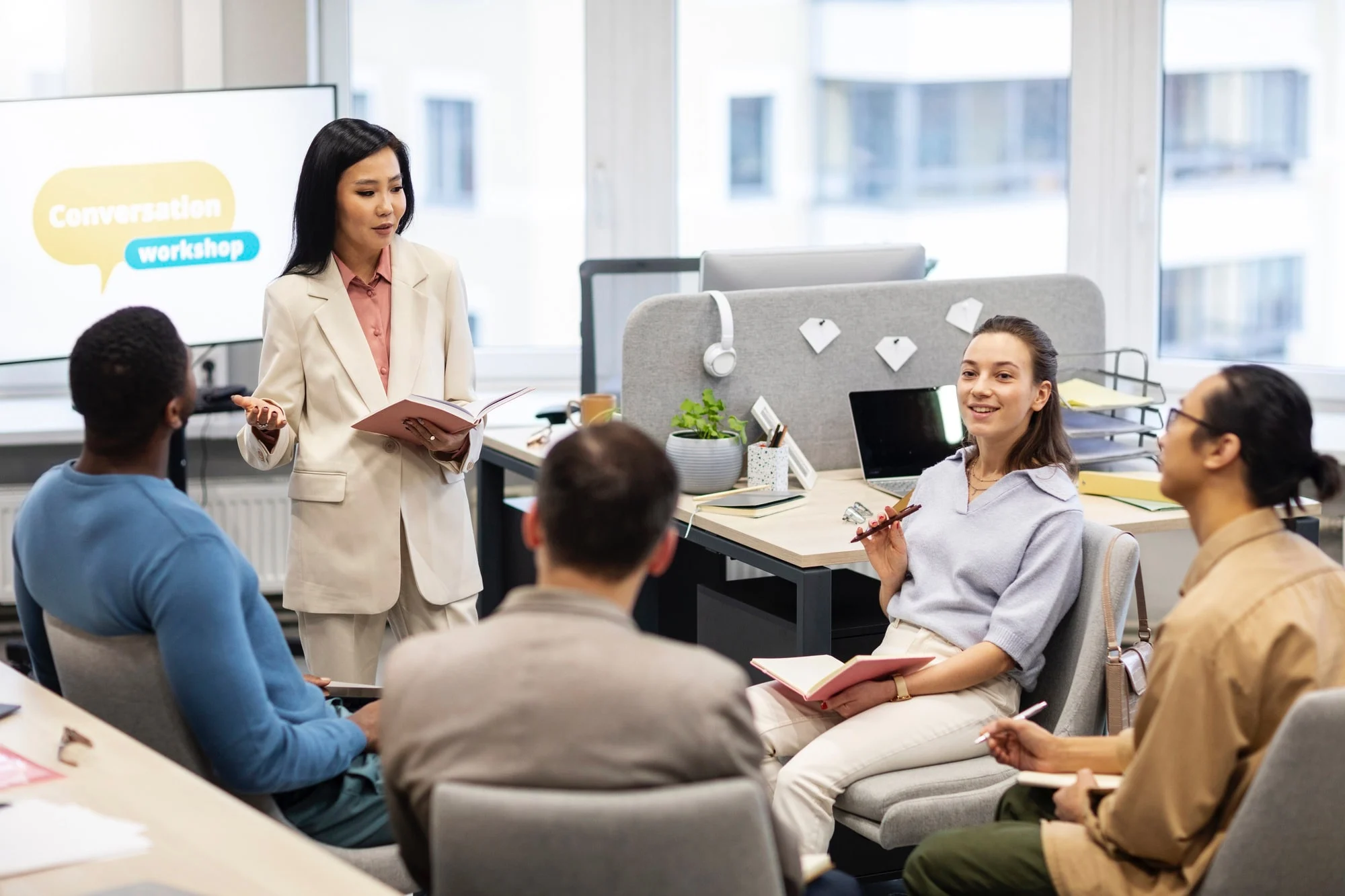 A woman leads a conversation workshop in an office, speaking to four seated participants who hold notebooks and pens. A presentation screen and desks are visible in the background.