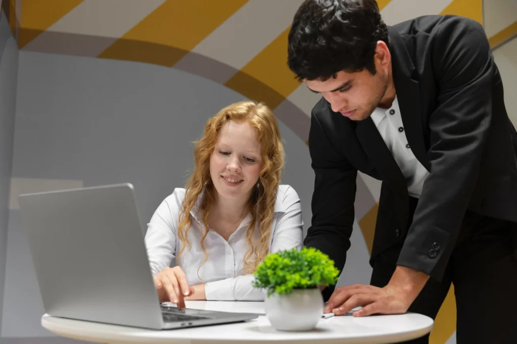 A woman sits at a table using a laptop while a man stands beside her, both looking at the screen. A small potted plant is on the table.