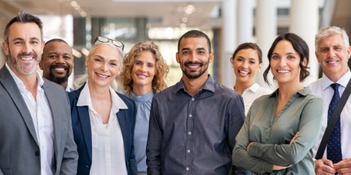 A group of eight professionally dressed people stand indoors, smiling and facing the camera in a well-lit office environment.