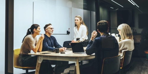 Five people sit and talk around a conference table in a modern office meeting room with laptops and notepads visible.