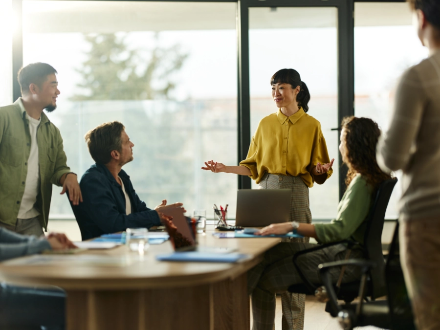 A woman stands and speaks to colleagues seated around a conference table in a modern office during a meeting.