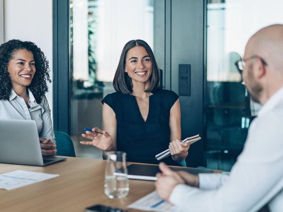 Three people have a discussion around a conference table, with documents, a laptop, and a glass of water visible. One woman is smiling and holding a notebook.