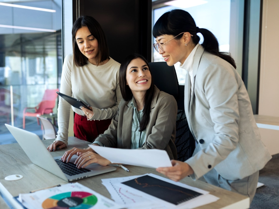 Three women in business attire collaborate at a desk with a laptop, tablet, and charts, engaging in discussion in a modern office setting.