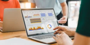 A person uses a laptop displaying various graphs and charts in a business analytics dashboard, with two other people standing nearby.