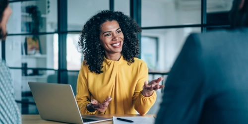 A woman in a yellow blouse sits at a desk with a laptop, smiling and gesturing while talking to two other people in an office setting.