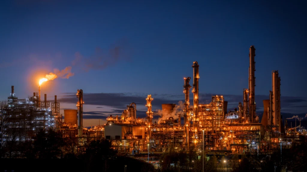 An illuminated oil refinery at dusk with multiple towers and a gas flare burning on the left side against a dark blue sky.