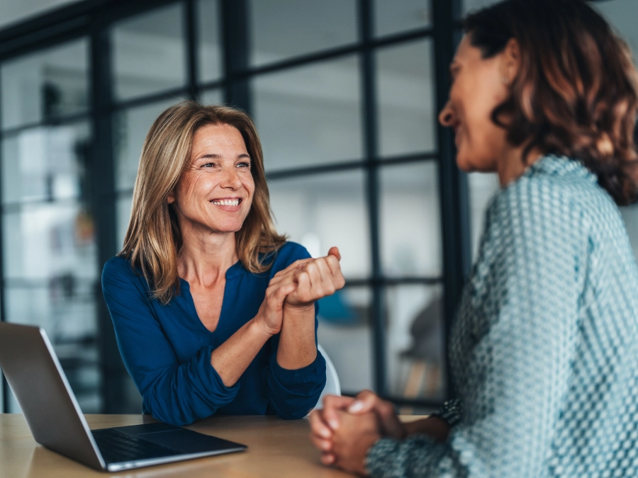 Two women are sitting at a desk having a conversation in an office setting, with a laptop open in front of them.