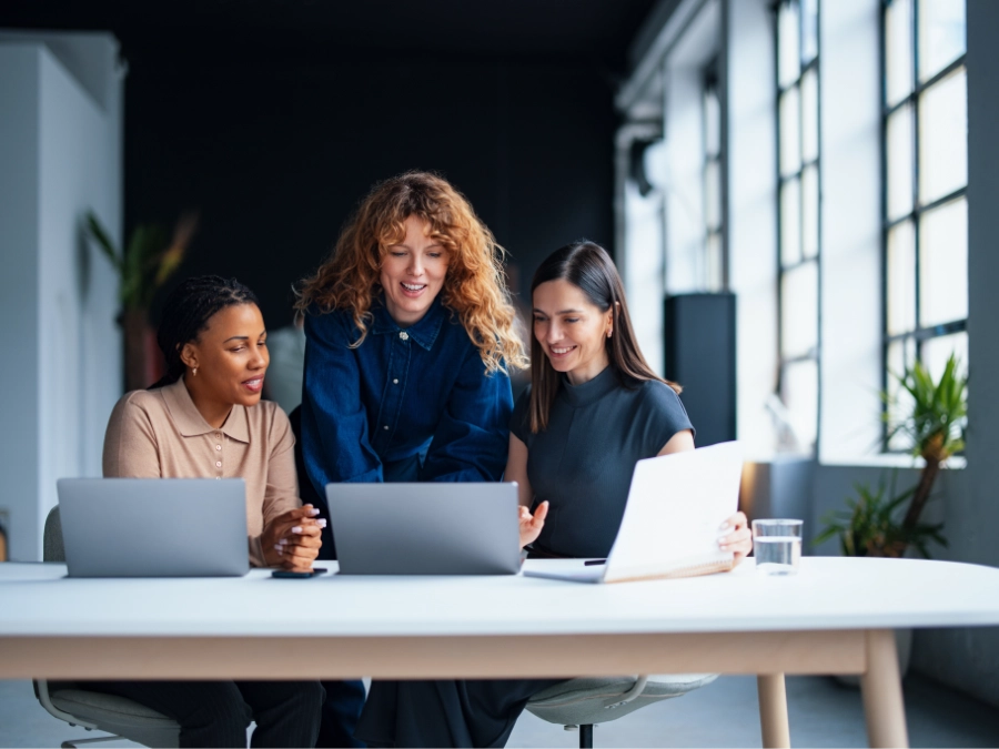 Three women sit at a table with laptops, collaborating and looking at a computer screen in a bright, modern office setting.